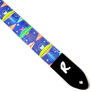 May include: A blue guitar strap with a repeating pattern of white cats in flying saucers with yellow, orange, and green accents. The strap has a black leather end with the letter "R" in white.