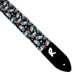 Peut inclure: Sangle de guitare noire avec le texte "TALK SHIT GET BIT" répété en blanc, rose et bleu sur fond noir. L'extrémité en cuir noir porte un "R" blanc et un trou pour l'attache.