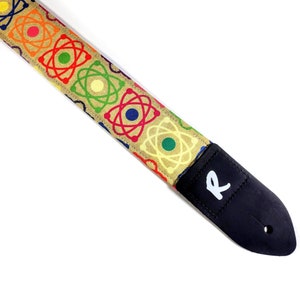 May include: A tan guitar strap with a colorful atomic symbol pattern. The strap has a black leather end with a white "R" logo.