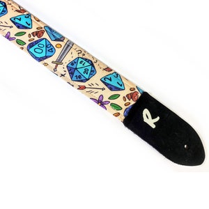 Op de afbeelding: Gitaarband met een beige patroon van blauwe dobbelstenen, zwaarden, pijlen en bloemen. De band heeft een zwart leren uiteinde met de letter "R" in het wit.