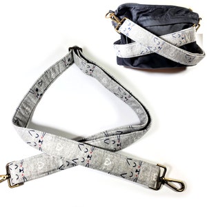 Könnte beinhalten: Ein verstellbarer Schultergurt in Grau und Weiß mit goldfarbener Hardware. Der Gurt hat ein sich wiederholendes Muster aus weißen Katzenköpfen mit rosa Nasen und blauen Augen.