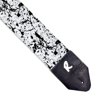 May include: White guitar strap with a black paint splatter design. The strap has a black leather end with a white "R" logo.