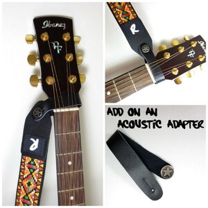 May include: A black guitar strap with a colorful woven pattern and a silver star. The strap has a black leather end with a silver star and the letter "R" on it. The strap is attached to an acoustic guitar with a brown wooden body and a gold headstock. The guitar has the word "Ibanez" on the headstock. The image also shows a black leather strap adapter with a silver star.