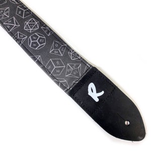 May include: A black guitar strap with a white 'R' on the end. The strap is covered in a pattern of white dice.