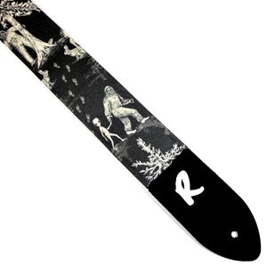 May include: Black guitar strap with a white illustration of Bigfoot, an alien, trees, and footprints. The strap has a white "R" logo and a hole for attachment.