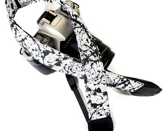 May include: A black and white camera strap with a splatter paint design. The strap is made of fabric and has a black adjustable strap.