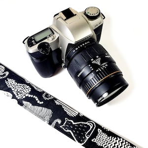 May include: A silver Pentax ZX-30 film camera with a black lens and a black and white patterned camera strap. The camera has a black lens with the text "SMC PENTAX-M 1:3.5-4.5 28-80mm" printed on it.