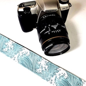 Könnte beinhalten: Ein blaues und weißes Kameraband mit Wellenmuster. Das Band ist an einer silbernen Pentax ZX-30 Kamera mit einem schwarzen Objektiv befestigt.