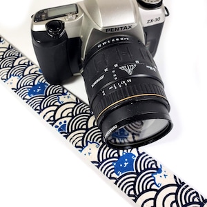 May include: A blue and white camera strap with a wave pattern and white cats. The strap is attached to a black and silver Pentax ZX-30 camera with a black lens.