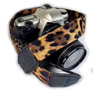 May include: A silver camera with a black and brown leopard print camera strap. The camera lens is a black "QUANTA RAY" 55mm lens.