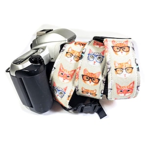 May include: A silver camera with a black strap featuring a repeating pattern of cats wearing glasses. The strap is made of fabric and has a black adjustable buckle.