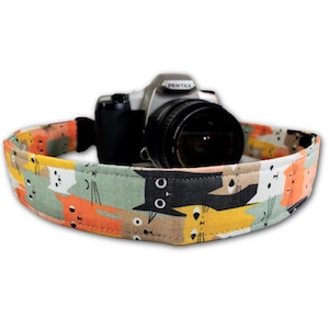 May include: A camera strap with a colorful cat pattern, featuring orange, yellow, white, and black cat faces on a green background. A silver and black Pentax camera rests inside the strap.