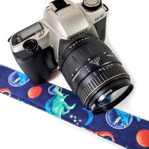 May include: A silver and black Pentax ZX-30 camera with a black lens and a blue strap. The strap features a pattern of planets, astronauts, and dinosaurs. The lens reads "QUANTAY 55mm".