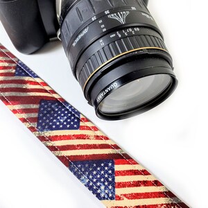 May include: A black camera lens with a silver ring and the text "QUANTARAY ASPHERICAl" printed on it. The lens is attached to a camera strap with a red, white, and blue American flag pattern.
