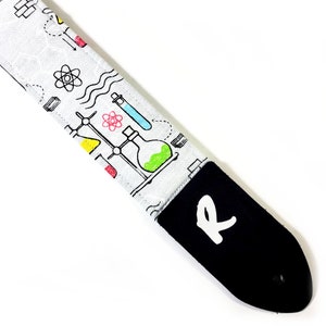 May include: White guitar strap with a black leather end. The strap is printed with colorful science-themed illustrations, including beakers, test tubes, and atoms.
