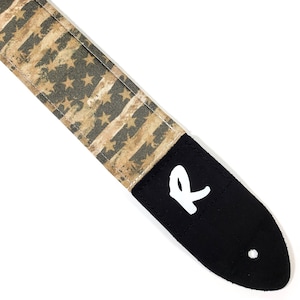 May include: A guitar strap with a camouflage American flag design. The strap features a black leather end with a white "R" logo and a single hole for attachment. The strap is made of durable fabric.