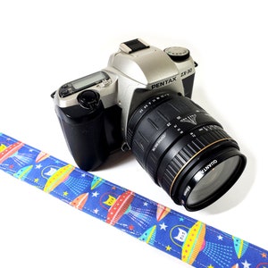 May include: A silver Pentax ZX-30 camera with a black lens and a blue camera strap with a cat and spaceship pattern.