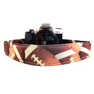 May include: A brown and white camera strap with a football pattern. The strap is made of fabric and has a stitched edge. A silver camera with a black lens is sitting on top of the strap.
