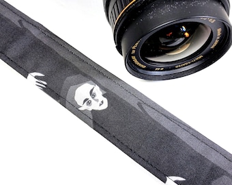 May include: A black camera strap with a white graphic print of a person's face. The strap is made of a textured fabric and has a stitched edge.