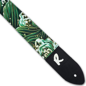 Op de afbeelding: Een gitaarband met een groen en goud golfpatroon. De band heeft een zwart leren uiteinde met de witte letter "R". De band staat diagonaal in het frame.