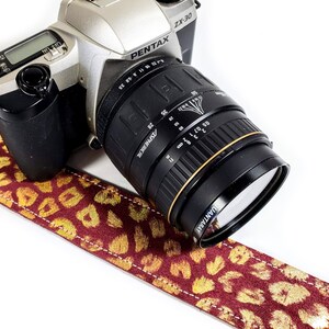 May include: A silver Pentax ZX-30 camera with a black lens attached. The camera has a black strap with a gold leopard print design.