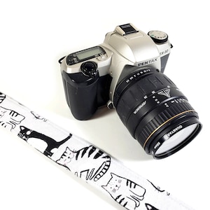 May include: A silver Pentax ZX-30 film camera with a black lens. The camera has a black and white cat-themed camera strap.
