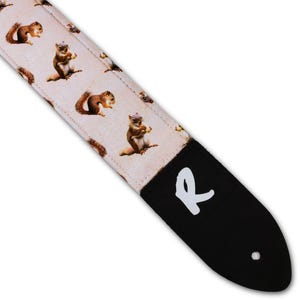 May include: A guitar strap with a cream-colored background featuring a repeating pattern of squirrels. The squirrels are in various poses, some holding nuts. The strap has a black leather end with a white "R" logo.