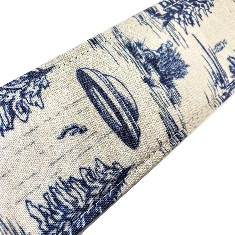 Pode incluir: Uma correia de guitarra bege com uma estampa azul e branca de um OVNI voando sobre uma floresta. A correia tem uma borda costurada.