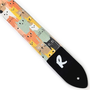 May include: A guitar strap with a repeating pattern of cartoon cats in shades of orange, green, yellow, and white. The strap has a black end piece with a white "R" logo and a hole for attachment.