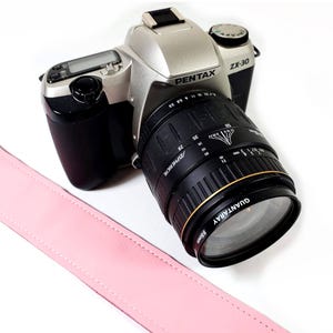 May include: A silver Pentax ZX-30 film camera with a black lens. The lens is a 35-70mm f/3.5-4.5 Aspherical lens. The camera has a pink camera strap.