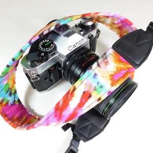 May include: A colourful tie-dye camera strap with black adjustable ends. The strap is attached to a silver Canon camera with a black lens.