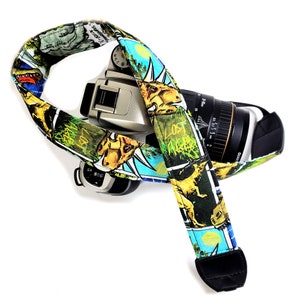 May include: A camera strap with a colorful dinosaur print. The strap is made of fabric and has a black adjustable strap. The strap is attached to a camera with a black lens.