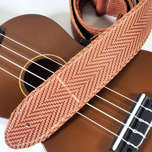 May include: A brown ukulele with white strings and a woven peach-colored strap. The strap has a herringbone pattern and white stitching. The ukulele is a musical instrument.