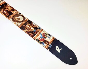 May include: A guitar strap with a brown and white fabric design featuring horses, saddles, and ropes. The strap has a black leather end with a white letter "R" on it.
