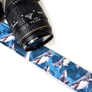 May include: A black camera lens with a blue and white shark-patterned camera strap. The lens has gold accents and the text "QUANTARAY 55mm". The strap is made of fabric and features a repeating pattern of sharks.