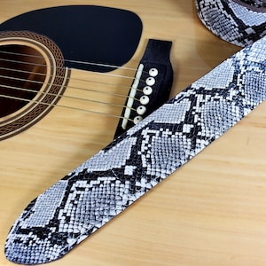 Peut inclure: Une sangle de guitare en peau de serpent noire et blanche avec un motif texturé. La sangle est attachée à une guitare acoustique.