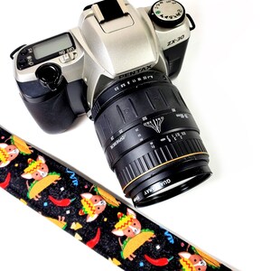 May include: A black camera strap with a colorful pattern of cartoon dogs wearing sombreros and holding tacos. The strap is attached to a silver Pentax ZX-30 camera with a black lens.