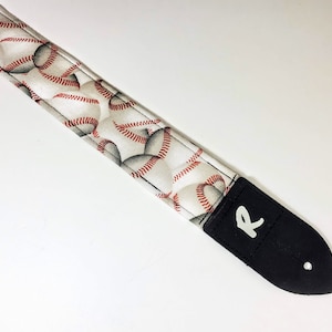 May include: White guitar strap with a baseball pattern and a black leather end with the letter "R" printed in white.