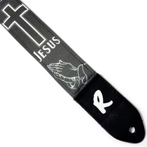 May include: A guitar strap with a gray background featuring a white cross, the word "JESUS", and praying hands. The strap has a black leather end with a white "R" and a hole for attachment.