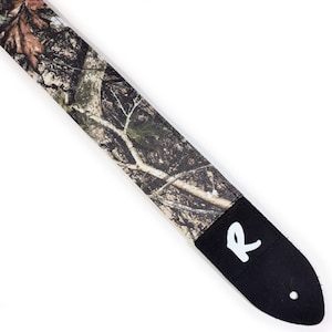 May include: A guitar strap with a camouflage pattern in shades of brown, green, and tan. The strap has a black leather end piece with a white "R" logo. The strap is angled diagonally across the frame.