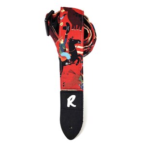 May include: Red guitar strap with a cowboy-themed pattern. The strap features illustrations of cowboys, horses, and other western imagery. The strap has a black leather end with the letter "R" in white.