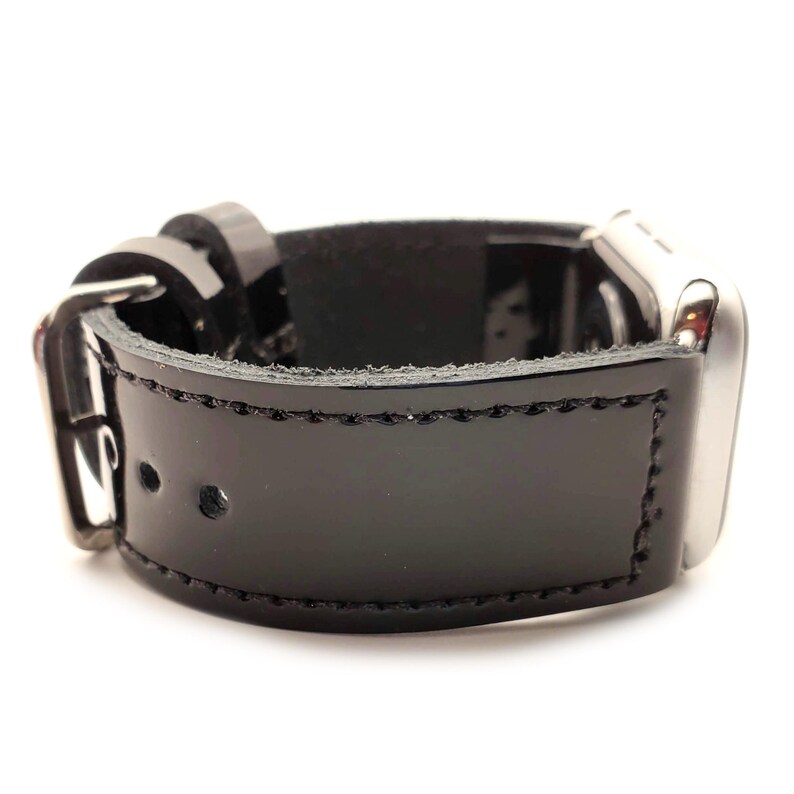 Puede incluir: Correa de reloj de cuero negro con hebilla y herrajes plateados. La correa es brillante y est&aacute; cosida en los bordes. La esfera del reloj es visible en el lado derecho.