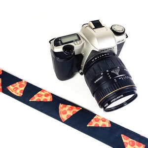 May include: A black and silver Pentax ZX30 camera with a pizza-patterned camera strap. The camera has a silver lens with a black lens cap.