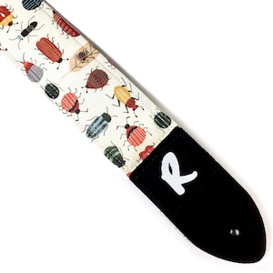 May include: A white guitar strap with a black leather end and a white letter "R" on the leather. The strap is covered in a pattern of colorful bugs.
