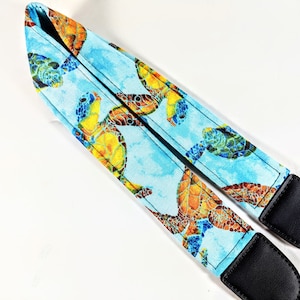 May include: A camera strap with a blue background and a pattern of colourful sea turtles. The strap is made of fabric and has black leather ends.