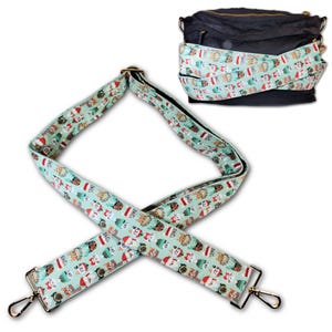 May include: A black crossbody bag with a patterned strap featuring a light blue background and a repeating design of cats wearing Santa hats and other festive attire. The strap has gold-colored metal hardware.