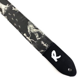 May include: Black and white guitar strap featuring a Bigfoot and alien design. The strap has a black suede end with a white "R" logo. The strap is made of a durable material and is designed for use with an electric guitar.
