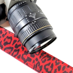 May include: A black camera lens with gold accents is partially visible, along with a red and black leopard print camera strap. The lens has markings and the word "QUANTARAY" is visible. The strap adds a stylish touch.