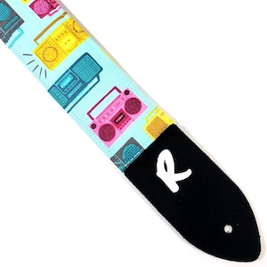 Puede incluir: Una correa de guitarra con fondo azul y un patrón de radios de colores. La correa tiene un extremo de cuero negro con un logotipo "R" blanco.