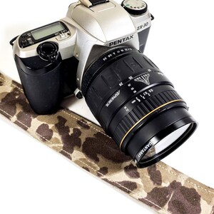 May include: A silver Pentax ZX-30 camera with a black lens. The camera is sitting on a camouflage-patterned strap.
